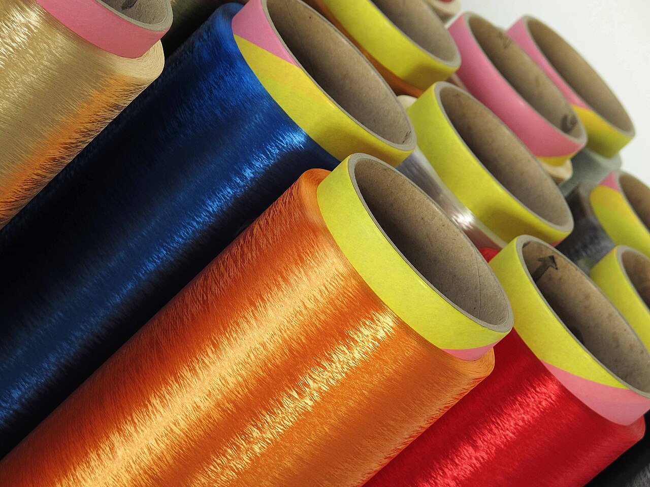 A close-up of packages of dyed filament yarn — glossy blue, orange, red and gold — wound on cardboard tubes and stacked on their sides.