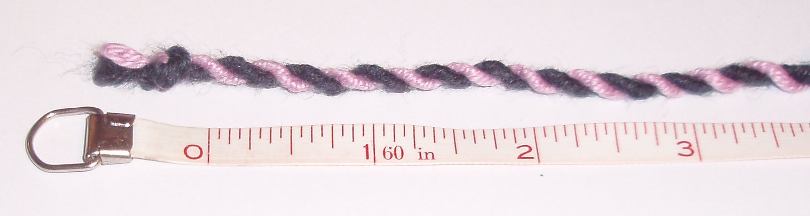 A length of two-ply yarn lying along a printed tape measure, photographed from directly above. One of the two strands is black and the other pink, so the two strands can be followed as they spiral round each other, and the individual turns can be counted against the inch marks on the tape.