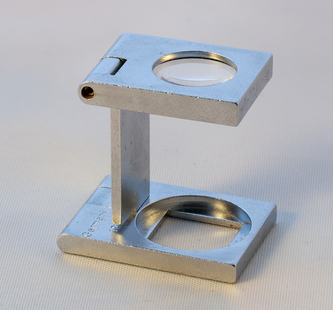 A folding metal linen tester standing open on a woven cloth, its magnifying lens on top and its square counting window in the base.