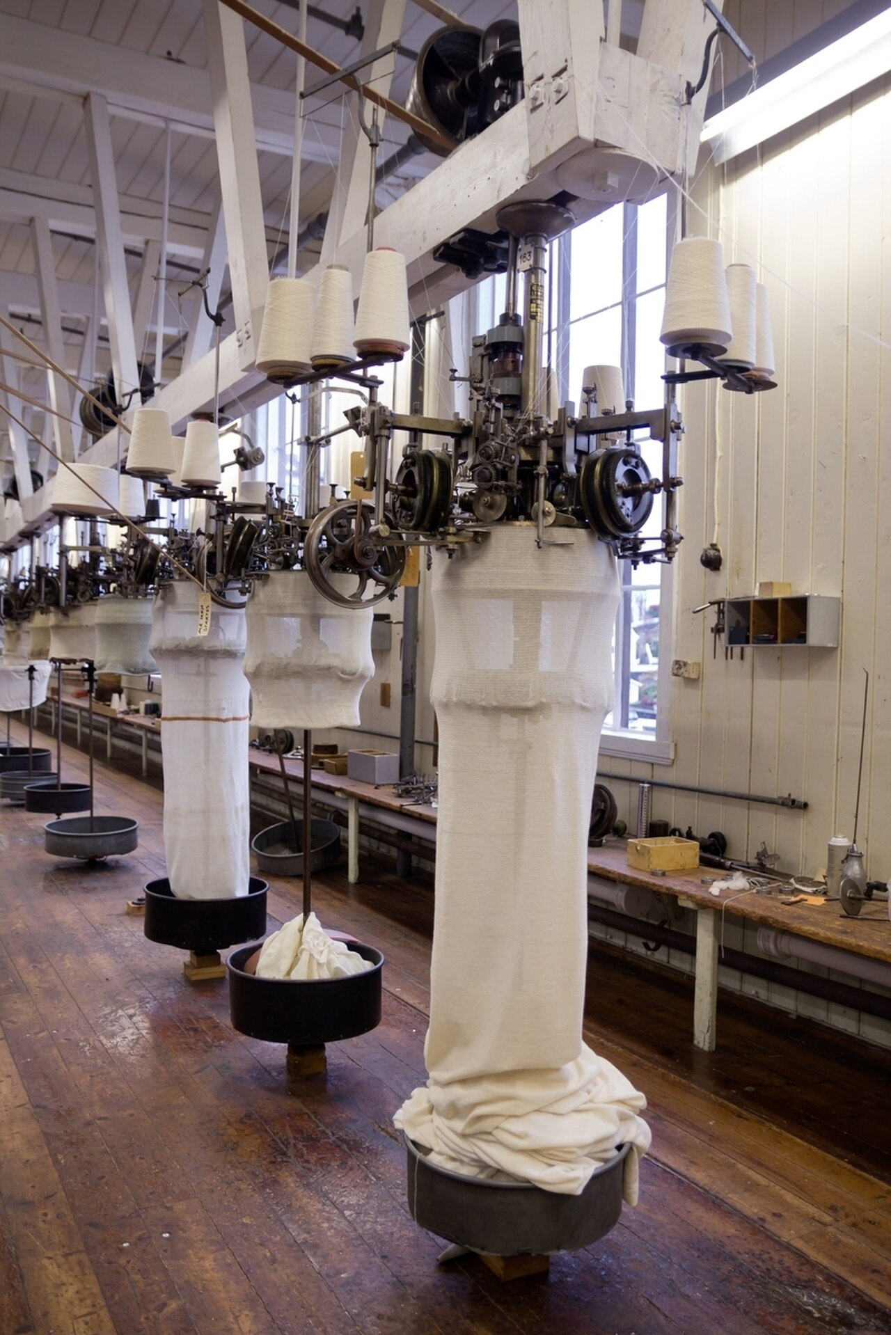 A row of circular knitting machines in a bright mill hall. Yarn cones sit above each machine, and a long white knitted tube hangs down from each cylinder into a round bin on the floor.