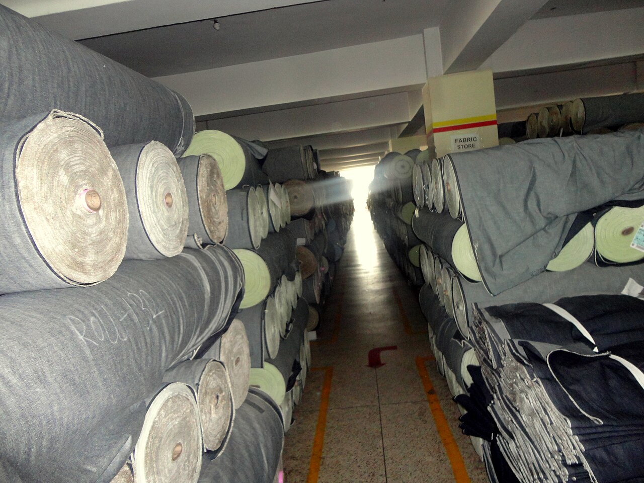 Looking down a narrow aisle in a factory fabric store, denim rolls stacked several high on both sides, a card sign overhead reading FABRIC STORE, and a roll number chalked on the end of one roll.