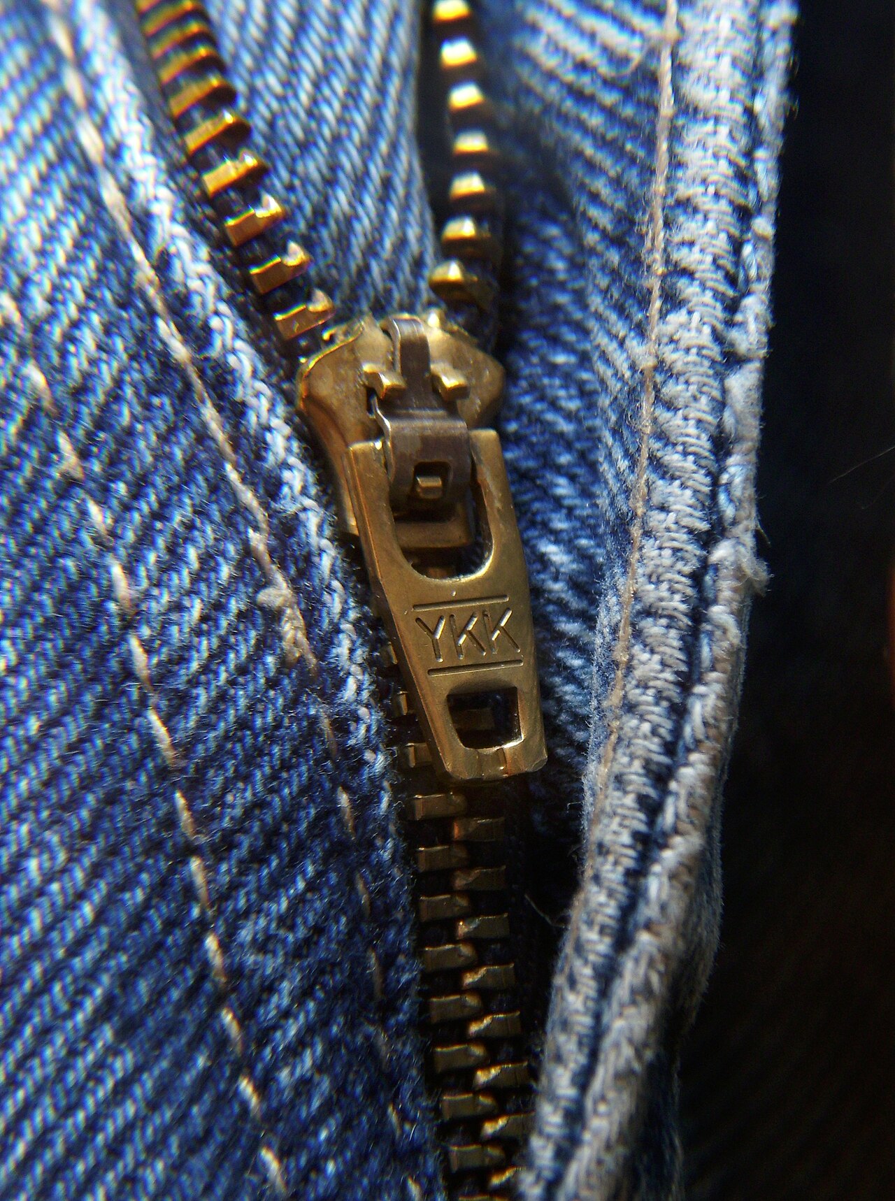 Close-up of a brass metal zip on blue denim. The slider is stamped YKK, and each tooth is clamped onto the tape one by one.