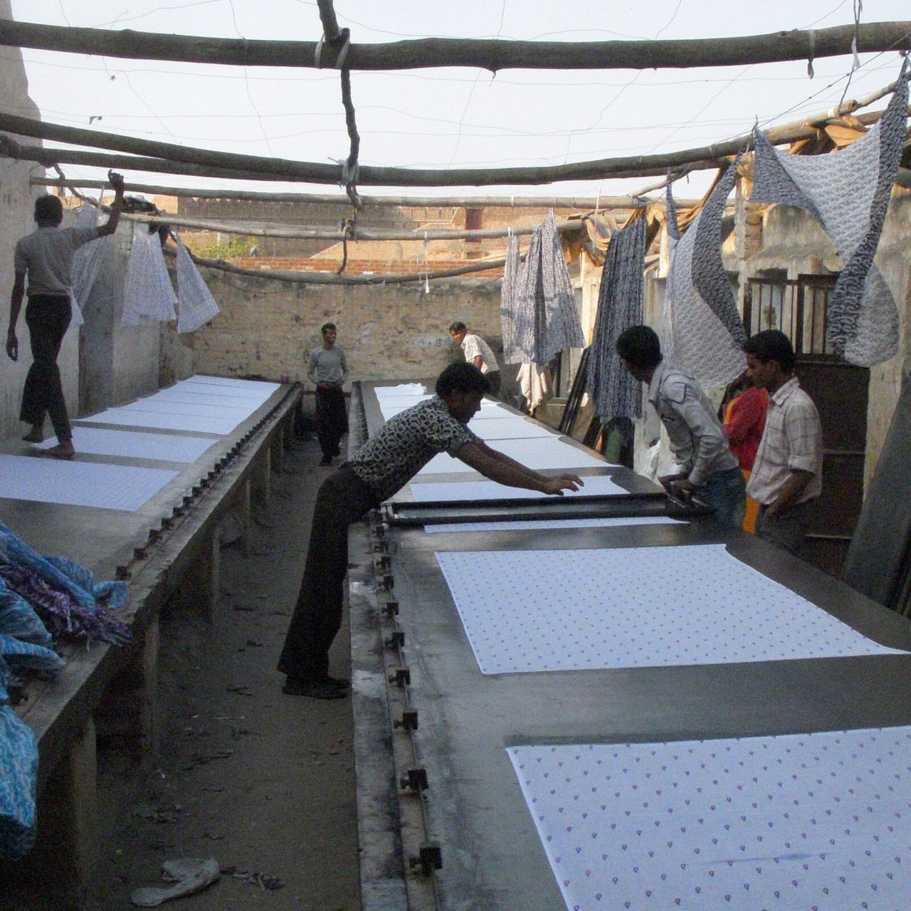 A long printing table under an open shelter, with white cloth pinned flat along it. Two men lift a flat screen frame clear of a freshly printed length. A small repeating motif has come off onto the cloth. Further down the same table an unprinted length waits. Behind them, printed lengths hang over lines to dry.