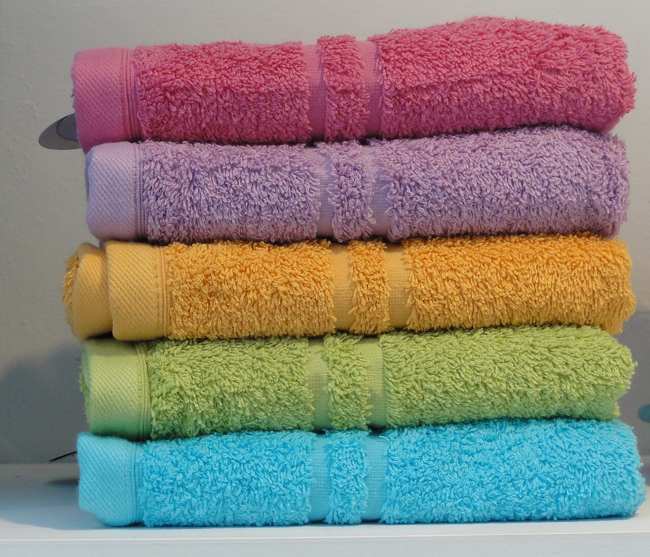 Five folded terry towels stacked in a pile — pink, purple, orange, green and blue — with the loop pile clearly visible on every fold.