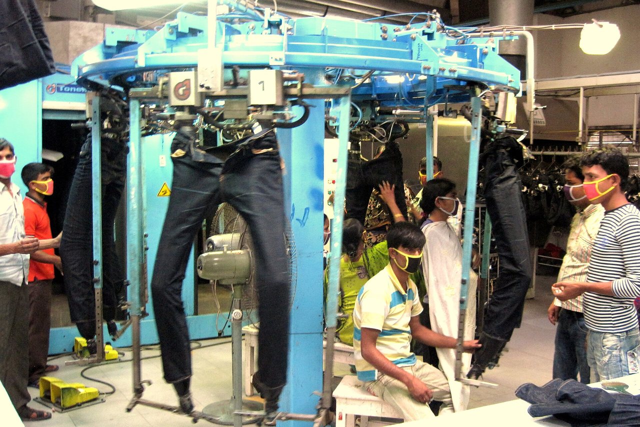 A denim finishing floor. Jeans are pulled over full-leg dummies mounted on a large blue rotating carousel, and operators in face masks work on the garments where they hang. More finished jeans hang on a rail behind the machine.
