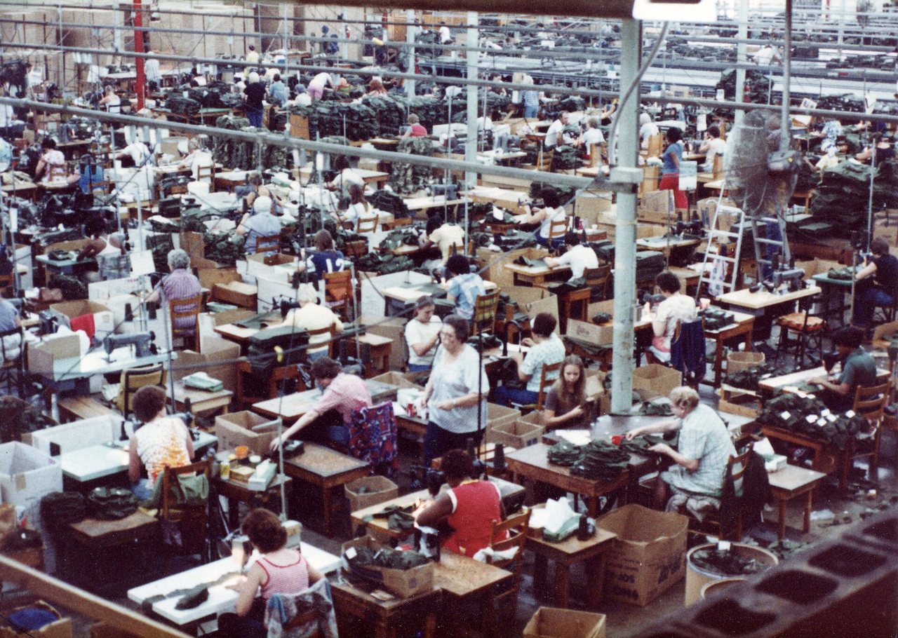 A large industrial sewing floor photographed from above. Several hundred sewing machines stand in long rows, an operator at most of them, with bins and cartons of part-finished garments beside each station and rails of work overhead.