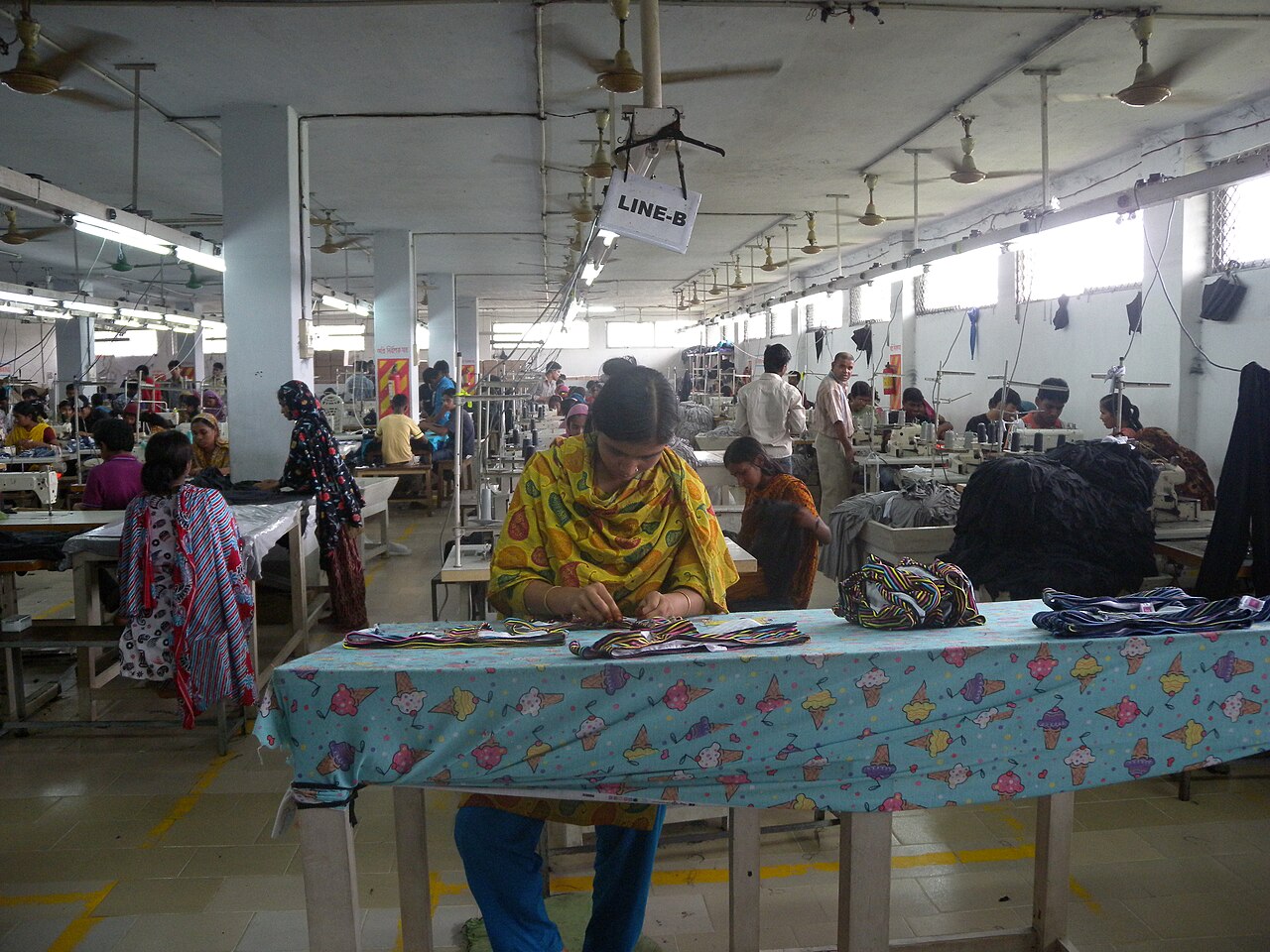 A sewing floor in a garment factory. Rows of machines run away from the camera under a hanging sign reading LINE-B, with operators at their stations and bundles of cut parts stacked on the tables.