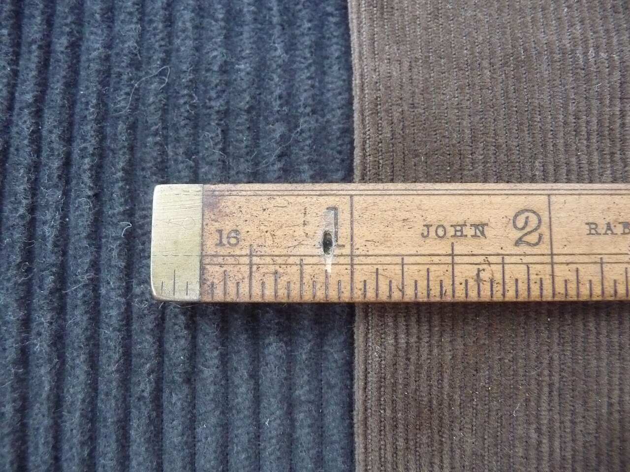 Two pieces of corduroy side by side under a wooden ruler. The cords run vertically, and one cloth has visibly wider cords than the other.