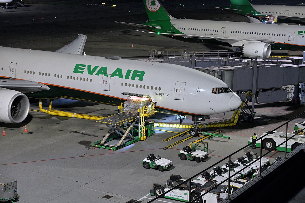 A shrink-wrapped pallet on a scissor loader, rising into the belly hold of a wide-body passenger jet at night, with tugs and dollies waiting on the apron.