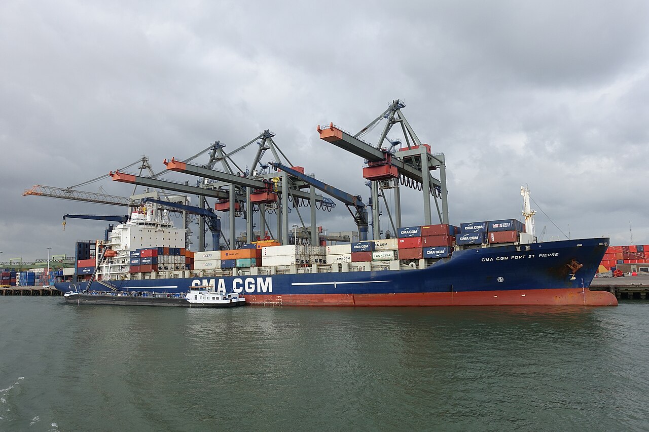 A container ship alongside a quay under four gantry cranes, with stacked containers on deck and on the quay behind.